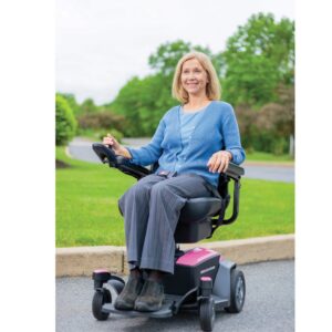 A woman in a blue sweater and gray pants sits on a Go Chair Power Wheelchair outdoors near a green lawn and trees.