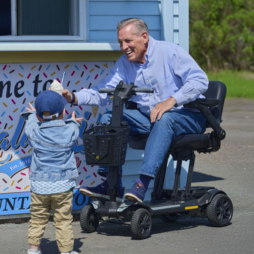 An older man in a mobility scooter hands something to a young child standing in front of him outside an ice cream shop on a sunny day.
