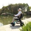 An older man sits by a lake in an Ally Power Wheelchair, holding a fishing rod, with trees and water in the background.