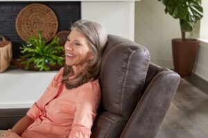 A woman with gray hair in a peach shirt smiles while sitting on the Titan Lift Chair, a brown recliner, in a plant-filled living room with a fireplace.