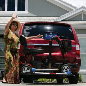A woman in a floral dress waves beside a red van equipped with the Harmar Heavy Duty Fusion Lift (AL300HD), which securely carries a mobility scooter mounted at the rear of the vehicle.