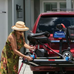A woman in a sun hat and floral dress uses the Harmar Heavy Duty Fusion Lift (AL300HD) to secure her mobility scooter to the rear carrier of a red SUV parked in a driveway.