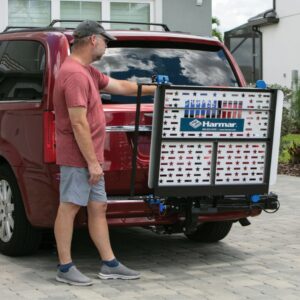 A man stands next to a red van, using a Harmar Heavy Duty Fusion Lift (AL300HD) attached to the rear, its platform decorated with stars and stripes.
