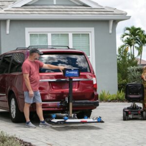 A man stands behind a red minivan in a residential driveway with a Harmar Heavy Duty Fusion Lift (AL300HD) attached, preparing to load a mobility scooter.