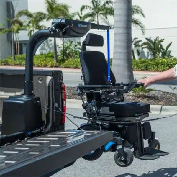 Using the Harmar Heavy-duty Power and Rotate Lift (AL425HD) attached to a vehicle, a person hoists a power wheelchair onto the open tailgate of a truck, set against a backdrop of palm trees and a road.