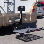 RV featuring a Harmar RV Lift (AL300RV) for an extra large scooter mounted at the rear, with a car partially visible in the background over a parking area.