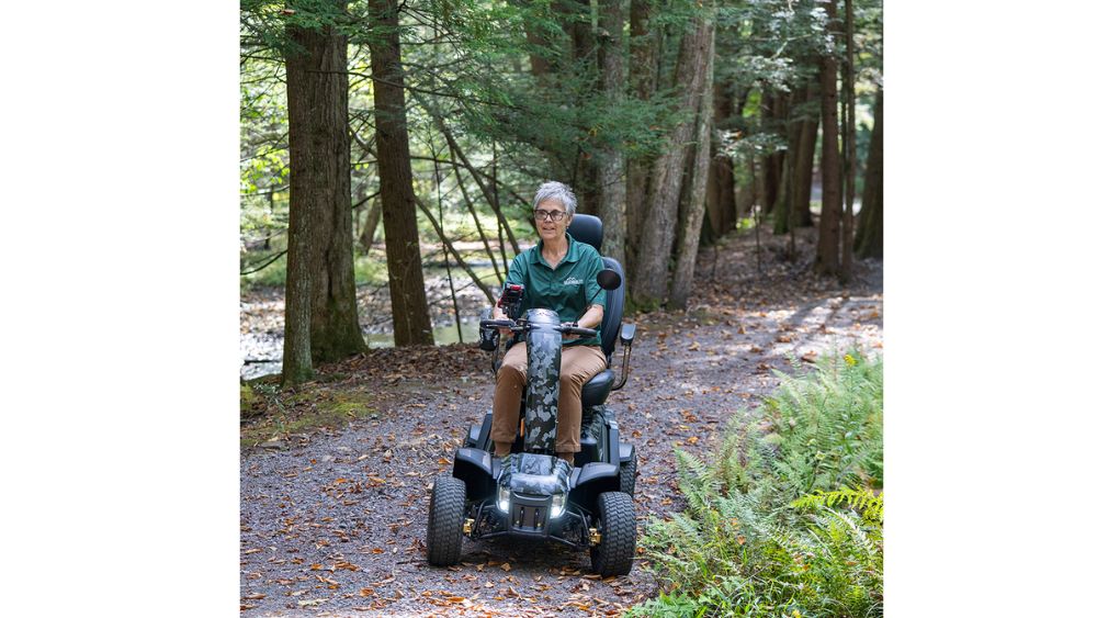 Riding the Baja™ Wrangler® 2 4-Wheel Scooter, a person travels along a dirt path surrounded by trees and ferns in a forested area.