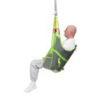 An older man sits in a gray and green Human Care Amputee Sling suspended from a ceiling lift, wearing white socks, gray sweatpants, and a white shirt.
