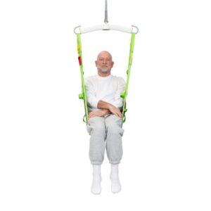 A man in a white shirt and gray sweatpants sits in a green Human Care Amputee Sling, suspended with his arms crossed and legs together, against a plain white background.