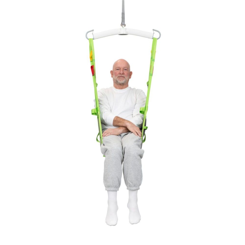 A man in a white shirt and gray sweatpants sits in a green Human Care Amputee Sling, suspended with his arms crossed and legs together, against a plain white background.