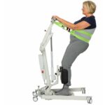 A woman stands on a patient lift with support straps around her torso, demonstrating assisted standing mobility equipment with the added safety of a Human Care Back Belt Sling.