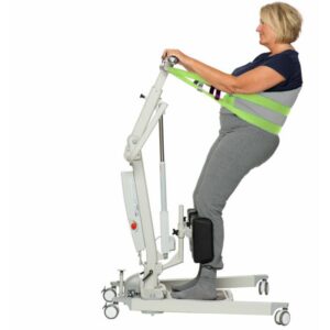 A woman stands on a patient lift with support straps around her torso, demonstrating assisted standing mobility equipment with the added safety of a Human Care Back Belt Sling.