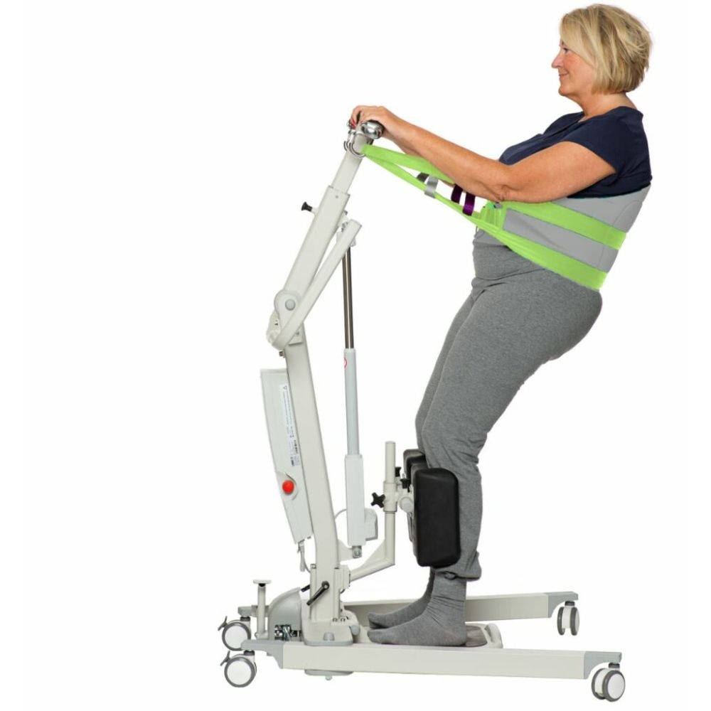 A woman stands on a patient lift with support straps around her torso, demonstrating assisted standing mobility equipment with the added safety of a Human Care Back Belt Sling.