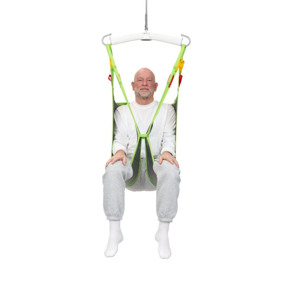 A man in white clothing sits in a green and yellow Human Care Basic Sling, suspended from a ceiling hoist against a plain white background.