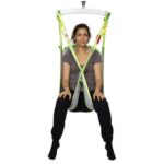 A woman sits in a green and gray Human Care Bath Sling-Polyester Net patient lift sling suspended from above, facing forward with her legs apart and hands in her lap.