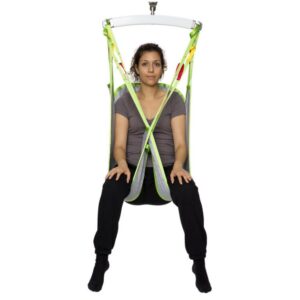 A woman sits in a green and gray Human Care Bath Sling-Polyester Net patient lift sling suspended from above, facing forward with her legs apart and hands in her lap.