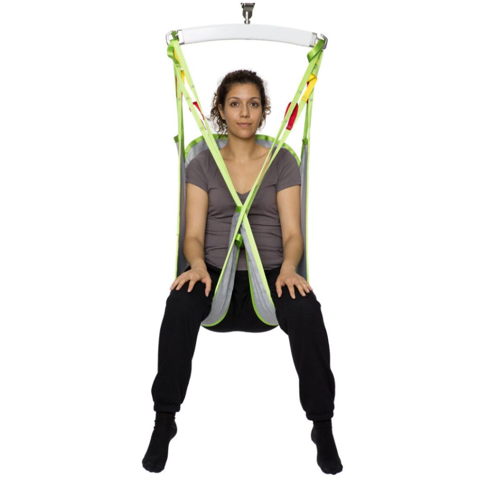 A woman sits in a green and gray Human Care Bath Sling-Polyester Net patient lift sling suspended from above, facing forward with her legs apart and hands in her lap.