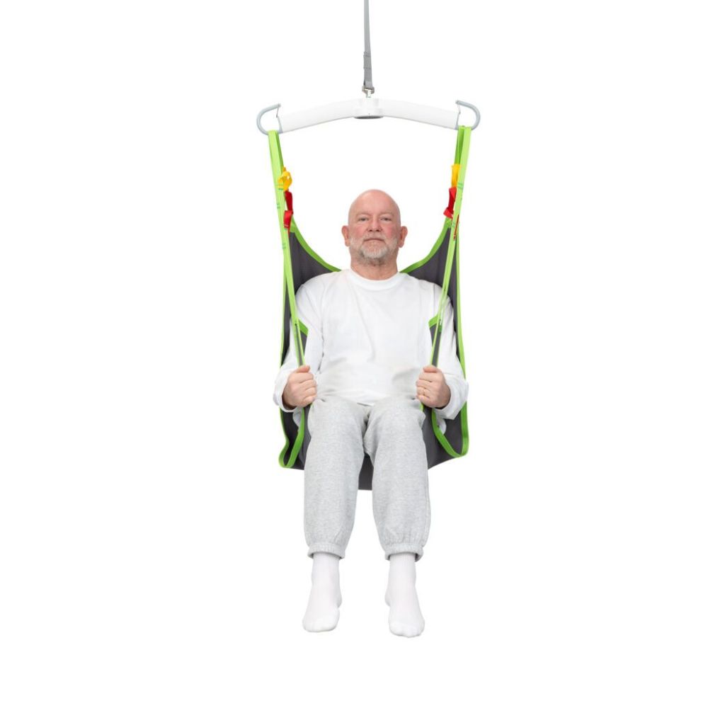 An older man in a white shirt and gray pants sits in a Human Care Comfort Sling, suspended from a ceiling hoist system, facing forward.