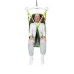 Older man sitting in a green and gray Human Care Full Body Sling patient lift sling, suspended off the ground, wearing a white shirt, gray sweatpants, and white socks.