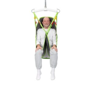 Older man sitting in a green and gray Human Care Full Body Sling patient lift sling, suspended off the ground, wearing a white shirt, gray sweatpants, and white socks.