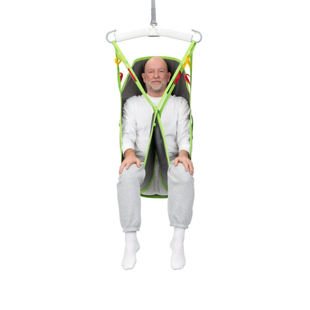 Older man sitting in a green and gray Human Care Full Body Sling patient lift sling, suspended off the ground, wearing a white shirt, gray sweatpants, and white socks.