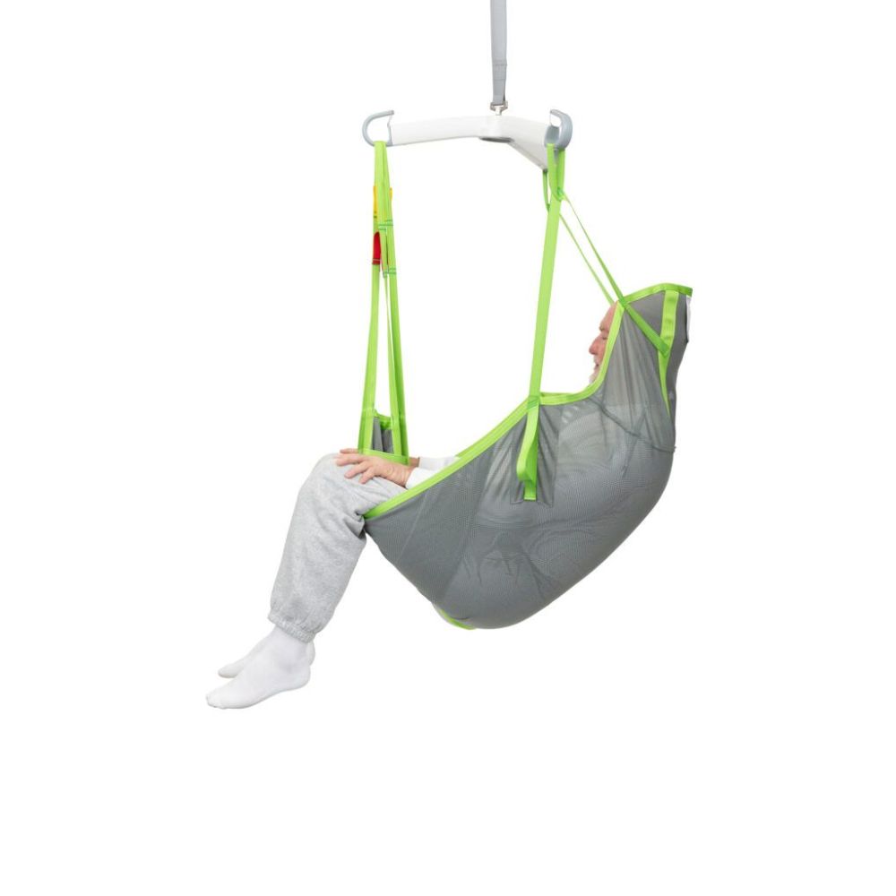 A person sits in a gray and green Human Care Full Body Sling, suspended from a white overhead bar, against a plain white background.