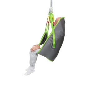 A person sits upright in a grey and green Human Care Full Body Sling patient lift sling, suspended by straps, with legs hanging and feet off the ground.