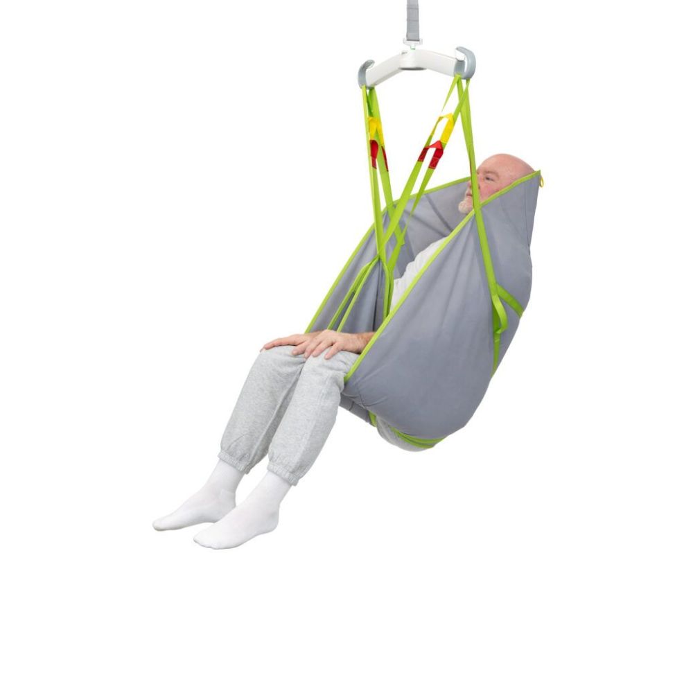 An older adult in gray pants and white socks sits comfortably in a Human Care General Purpose Sling, supported by a gray and green patient lift sling and suspended by a ceiling lift system against a white background.