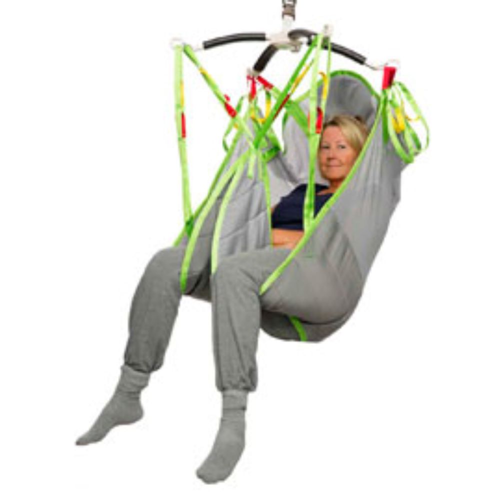 An adult woman is sitting in a gray Human Care Highback Plusline Sling with green straps, suspended from a mechanical patient lift, against a white background.