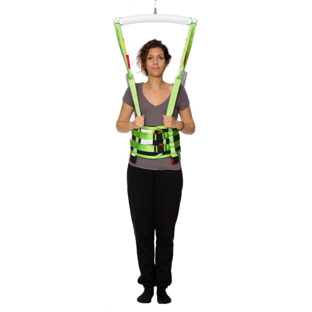 A woman stands upright wearing a green Human Care Multi Sling patient transfer harness attached to overhead straps, demonstrating a medical lifting device.