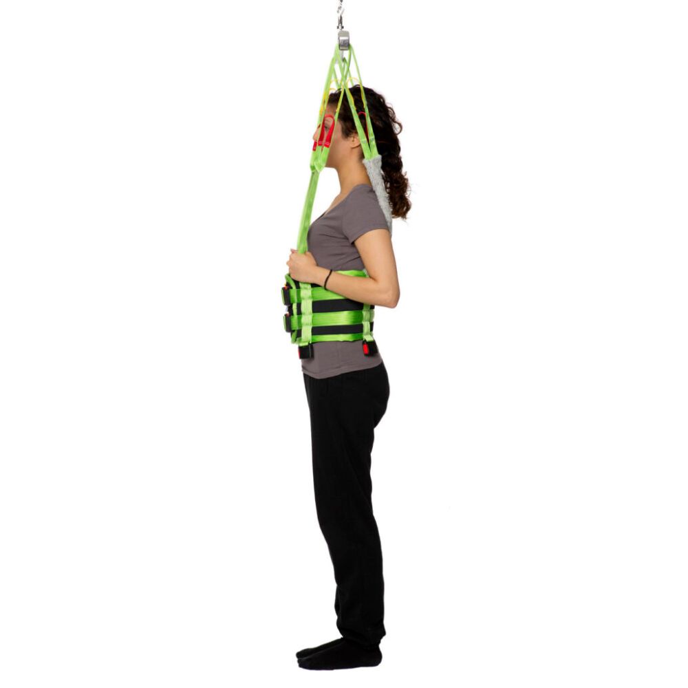 Person standing upright wearing a green Human Care Multi Sling patient lift harness attached to an overhead support, demonstrating a medical lifting aid.