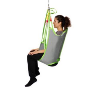 A woman sits upright in a grey and green Human Care Sit Sling- Polyester, suspended from a ceiling hoist, demonstrating its use for assisted transfer.