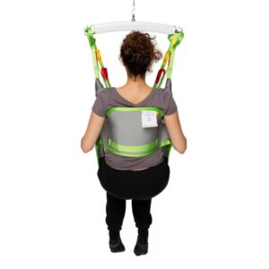 A person with curly hair sits in a gray and green Human Care Toileting Sling, suspended from a white bar, facing away from the camera.