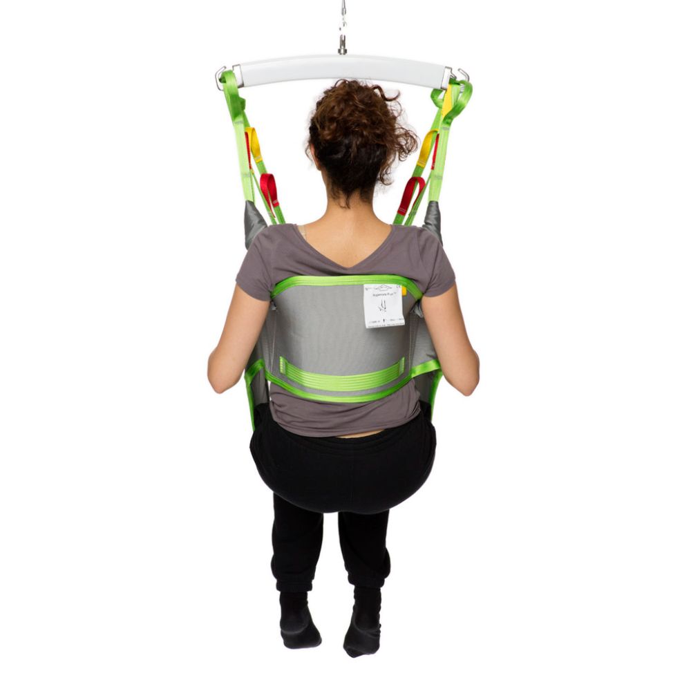A person with curly hair sits in a gray and green Human Care Toileting Sling, suspended from a white bar, facing away from the camera.