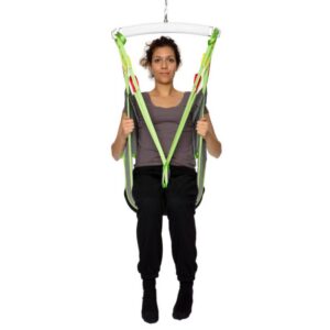 A person sits in a Human Care Toileting Sling with green and yellow straps, suspended above the ground against a white background.