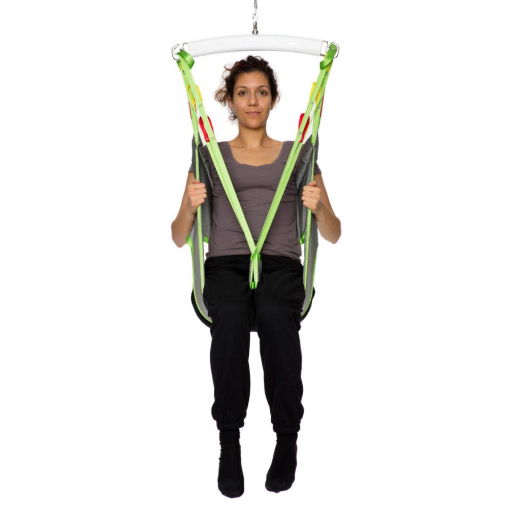 A person sits in a Human Care Toileting Sling with green and yellow straps, suspended above the ground against a white background.