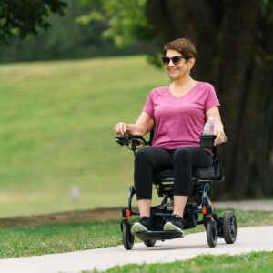 A woman wearing sunglasses and a pink shirt rides a Jazzy Carbon 27 Power Wheelchair along a park path, holding a water bottle and smiling.