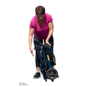 An older woman in athletic clothing bends down to lift or move a folded, blue Jazzy Carbon 27 Power Wheelchair with wheels.