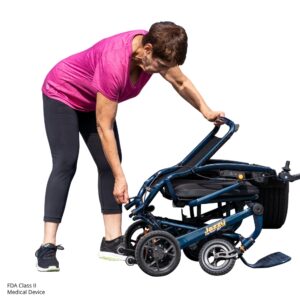An older woman in athletic wear bends down to fold a Jazzy Carbon 27 Power Wheelchair with a blue frame.