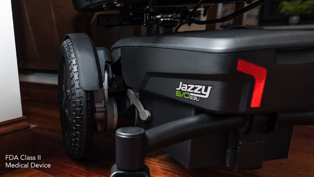 Close-up of the rear wheel and frame of a Jazzy EVO 613 Power Wheelchair on a wooden floor, with “FDA Class II Medical Device” text in the lower left corner.