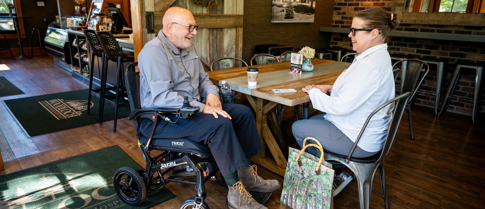An older man in a wheelchair and a woman sit at a wooden table in a café, conversing over coffee. A floral bag rests on the floor near the woman.