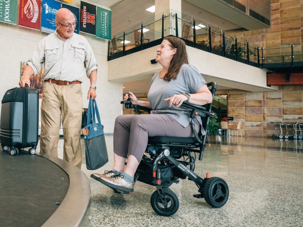 A woman in a motorized wheelchair talks to a man carrying luggage in an airport terminal.