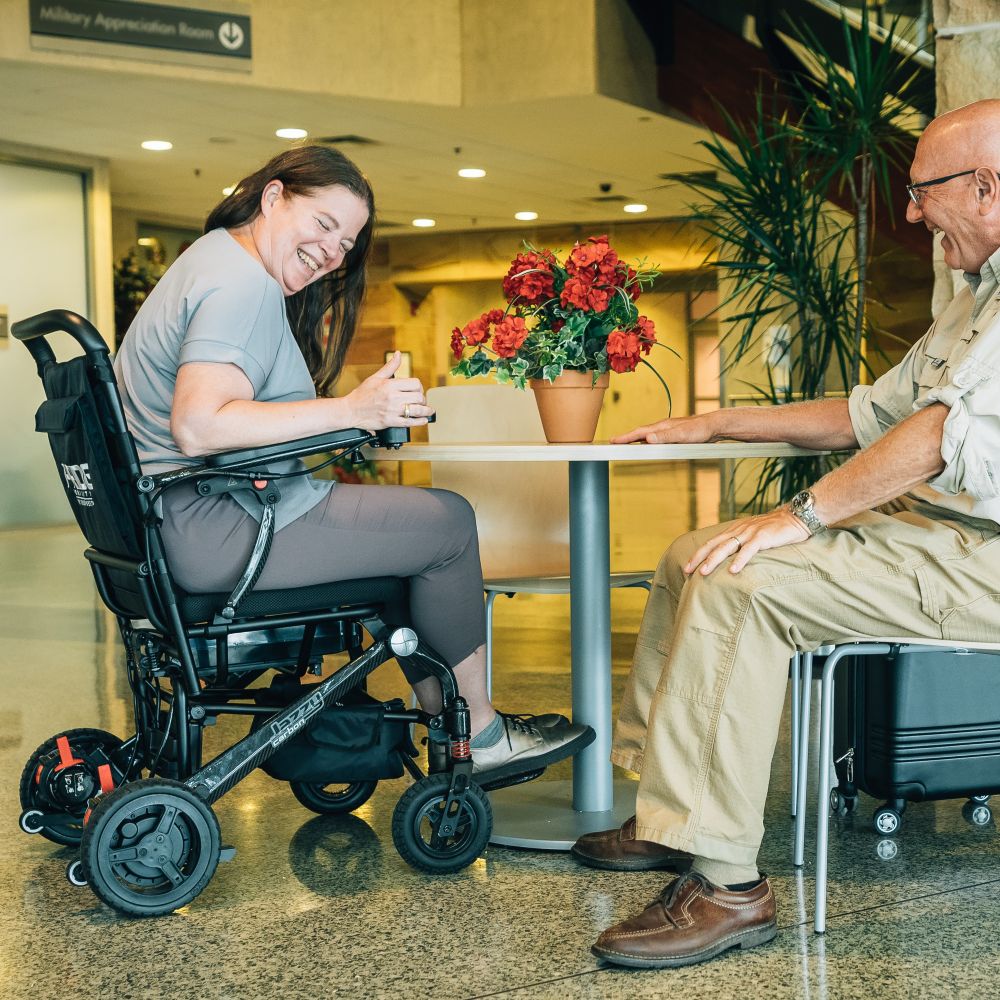 A woman in a wheelchair smiles while sitting at a table with an older man in a public indoor setting with a potted plant between them.