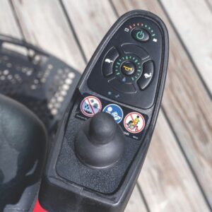 Close-up of a Jazzy Elite HD Power Wheelchair control panel featuring a joystick, power button, horn, battery indicator, and warning stickers. Wooden planks are visible in the background.