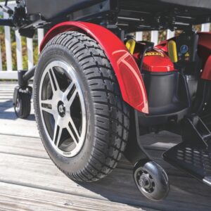 Close-up of a large rear wheel and a small anti-tip caster on a red Jazzy Elite HD Power Wheelchair, parked on a wooden deck.