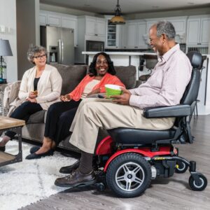 Three adults are gathered in a living room, with one man seated in a Jazzy Elite HD Power Wheelchair holding a book and conversing with two women who are sitting together on the couch.