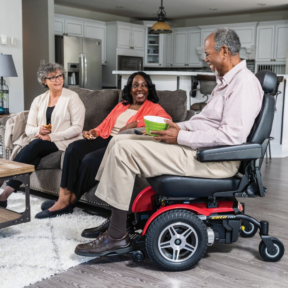 Three adults are gathered in a living room, with one man seated in a Jazzy Elite HD Power Wheelchair holding a book and conversing with two women who are sitting together on the couch.