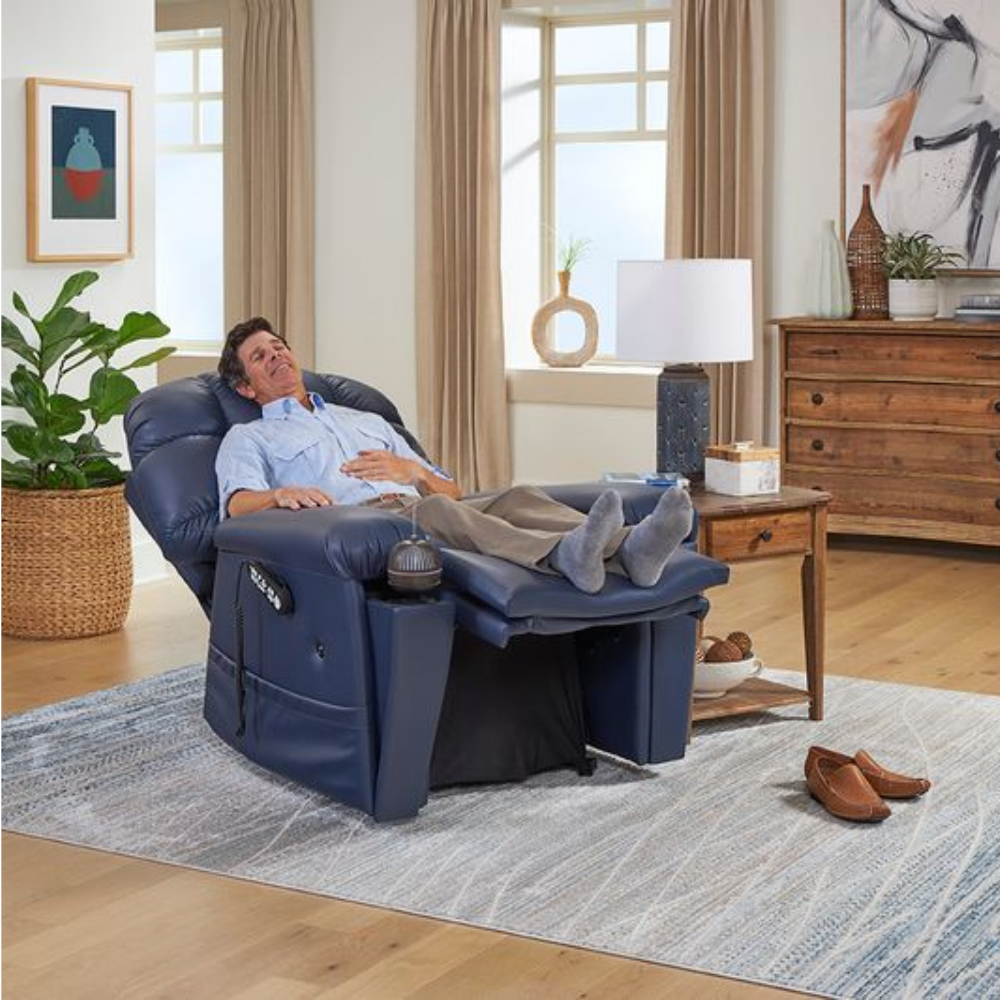 A man reclines in a Cloud Supreme with Twilight Lift Chair, his feet up, in a living room with wooden floors, a rug, plants, and a side table with a lamp, basking in comfort and serenity.