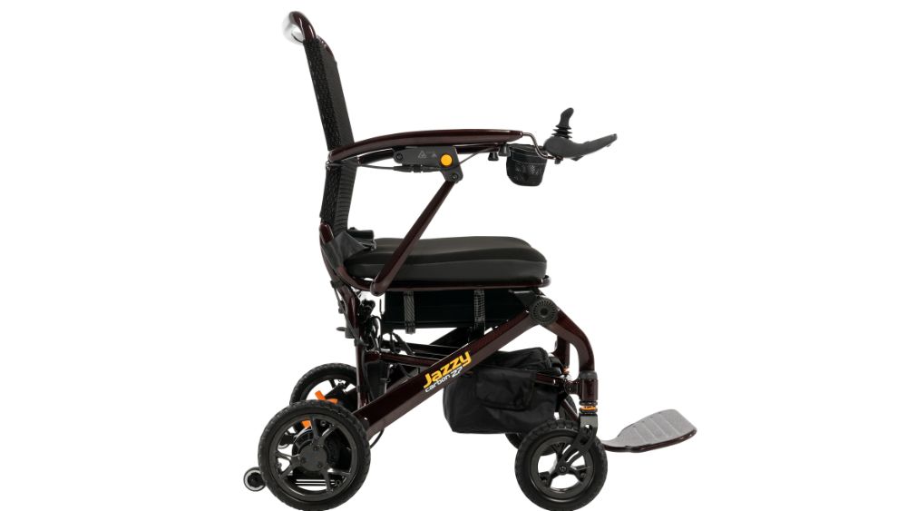 Side view of the Jazzy Carbon 27 Power Wheelchair—a lightweight, foldable model with a black seat, armrests, controller joystick, compact wheels, and “Jazzy” branding on the frame.