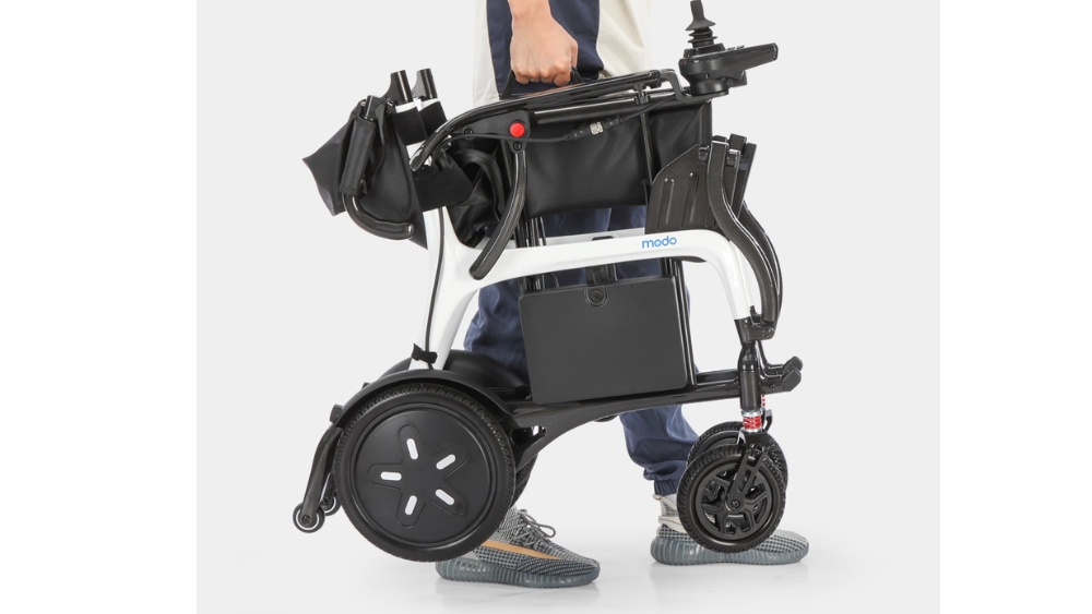 A person stands holding a folded Modo Breeze electric wheelchair in one hand, showcasing its compact and portable design.